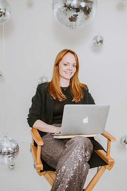 christine coughlin sits in a directors chair with a laptop on her lap smiling at the camera